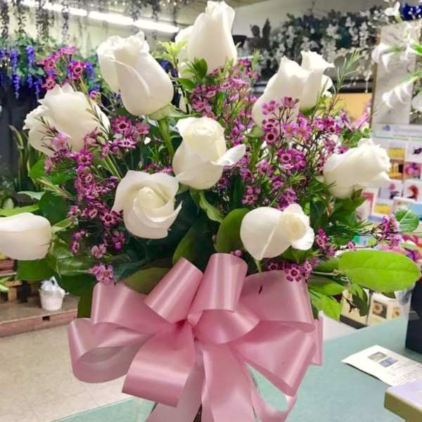 White roses and purple filler flowers in a glass vase with a pink bow