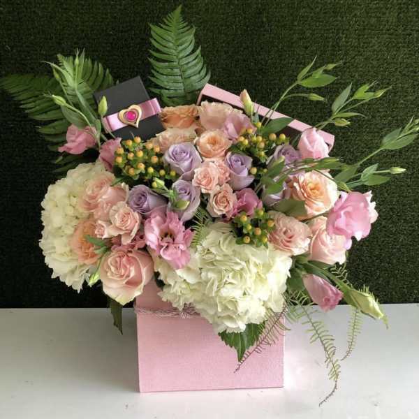 Pastel roses, hydrangeas, and lisianthus in a pink box with a small gift box among the blooms