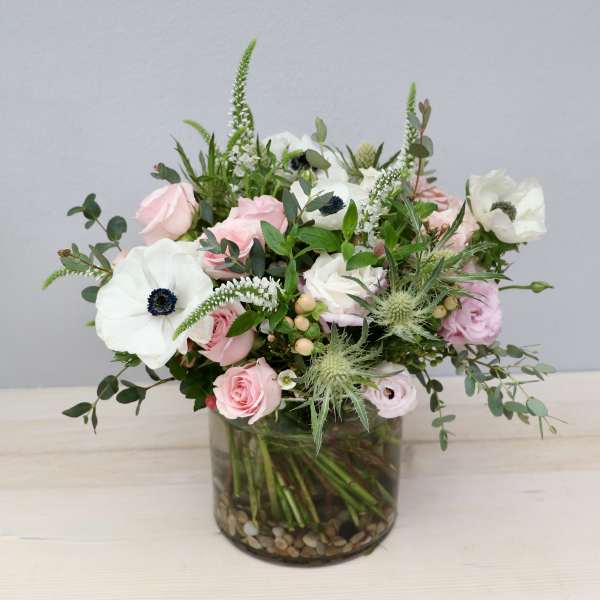Pastel pink roses and white anemones arranged in a glass cylinder vase with stones.
