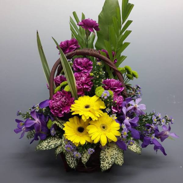 Basket arrangement with yellow gerbera daisies, purple flowers, and tropical leaves