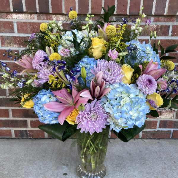 Mixed bouquet of blue hydrangeas, pink lilies, and yellow roses in a glass vase