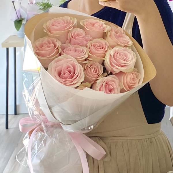 Handheld bouquet of light pink roses wrapped in white paper and cellophane with a pink ribbon.
