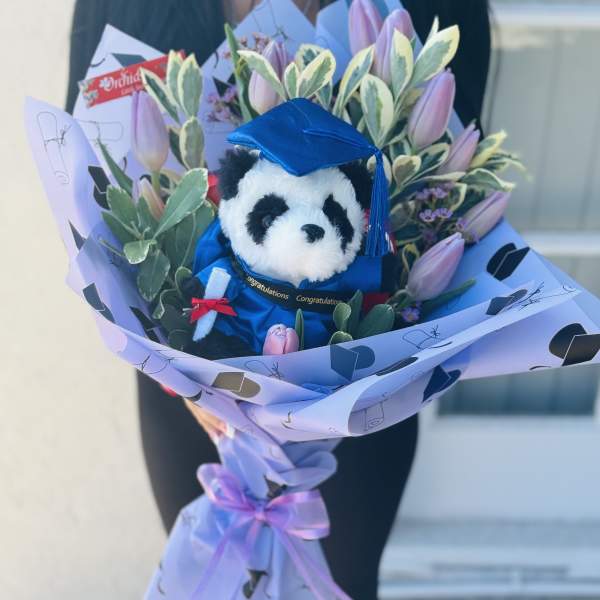 Bouquet with a plush panda in a blue graduation cap surrounded by pink tulips
