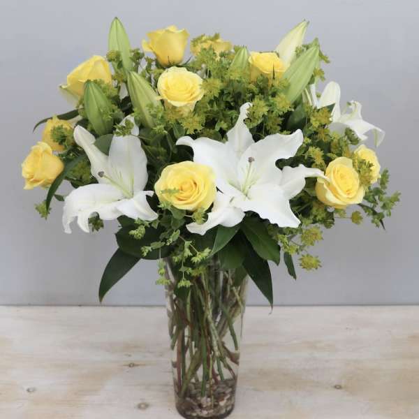 Tall glass vase of yellow roses and white lilies with green filler flowers