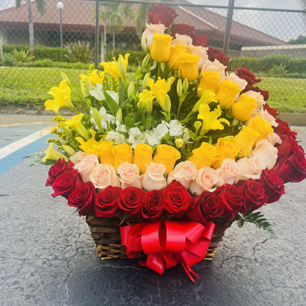 Basket arrangement of red, yellow, and blush roses with lilies and a red bow