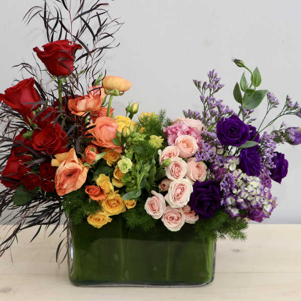 Low rectangular arrangement of red, yellow, peach, and purple flowers in a glass vase.