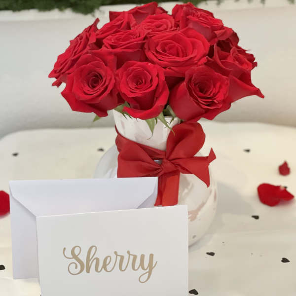Red roses in a white vase with a red ribbon and a name card