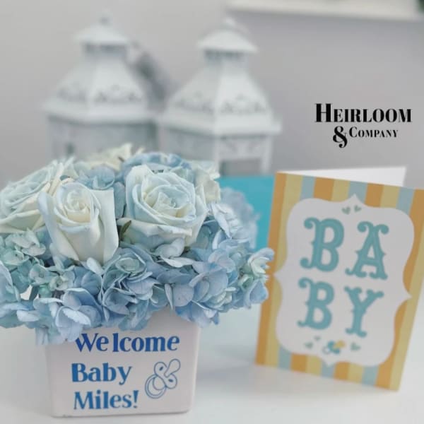 Blue and white flower arrangement in a white container with a baby card