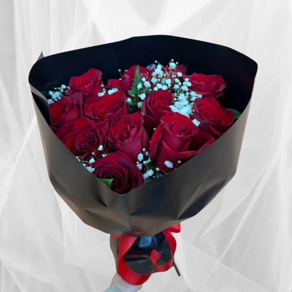 Handheld bouquet of red roses with white filler wrapped in black paper and red ribbon.