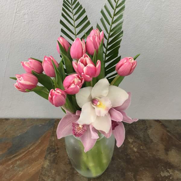 Pink tulips and orchids arranged in a glass vase with tall palm fronds.