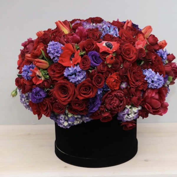 Low dome of red roses and purple blooms arranged in a black hatbox-style container