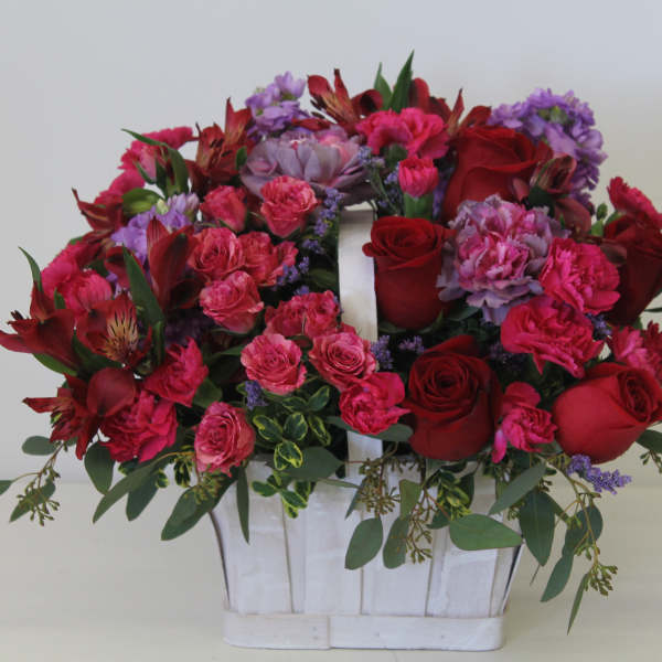 Bright basket arrangement of red roses, pink carnations, and purple blooms in a white wooden handled container