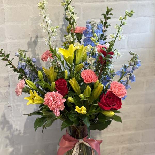 Tall mixed bouquet of yellow lilies, red roses, pink carnations, and blue delphinium in a glass vase with ribbon