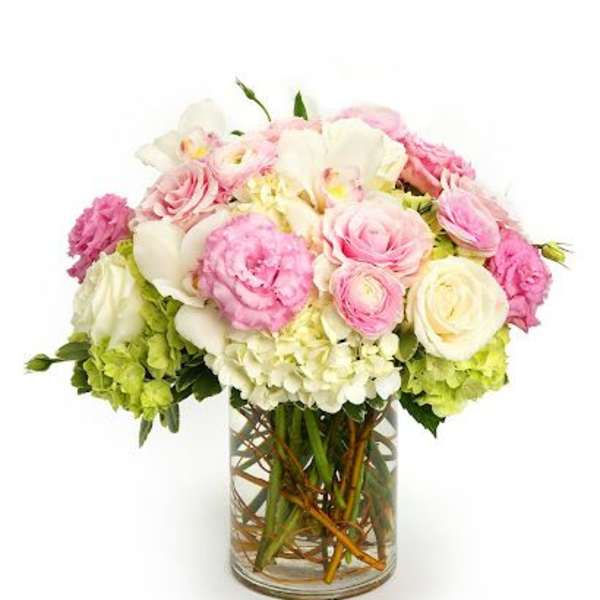 Round arrangement of pink and white roses, hydrangeas, and other blooms in a clear glass cylinder vase