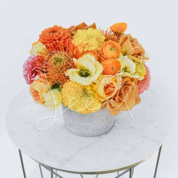 Low arrangement of orange, yellow, and peach flowers in a white pot on a round marble table.
