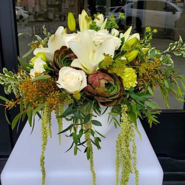 Low white bowl arrangement with white lilies, roses, succulents, and trailing green accents