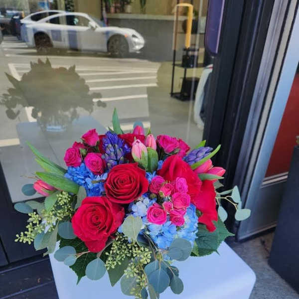 Round arrangement of red roses, pink blooms, blue hydrangea and purple flowers in a low vase