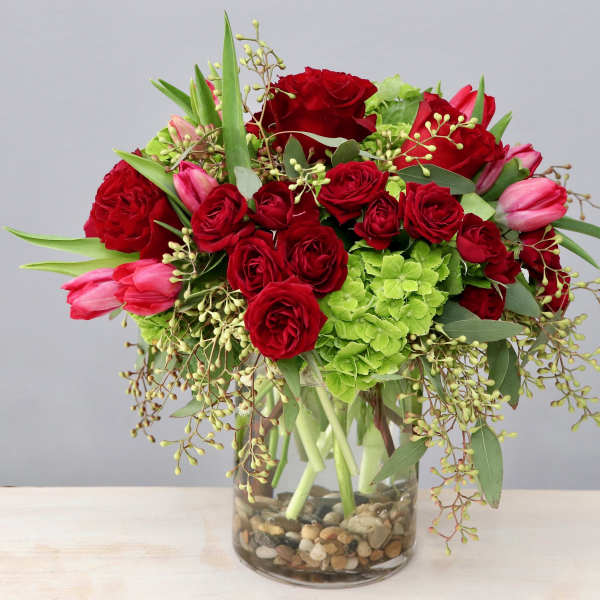 Arrangement of red roses, pink tulips, and green hydrangeas in a clear glass vase with river rocks