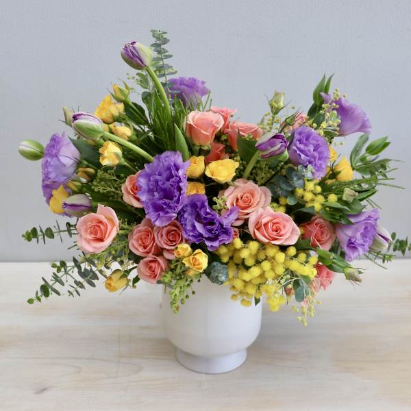 Colorful arrangement of purple lisianthus, peach roses, and yellow blooms in a white ceramic vase