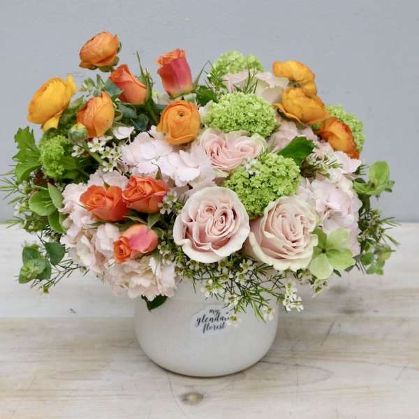Low arrangement of peach roses, orange ranunculus, and green blooms in a white ceramic pot.