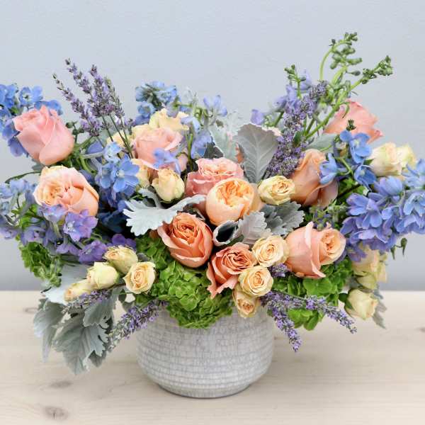 Low arrangement of peach roses and blue flowers in a white ceramic bowl vase