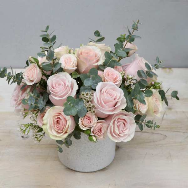 Low arrangement of pale pink roses and spray roses with eucalyptus in a white ceramic vase