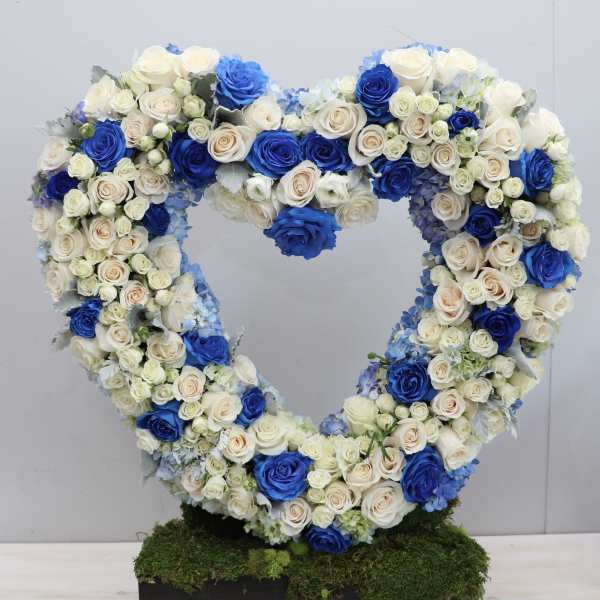 Heart-shaped standing wreath of blue and ivory roses on a moss-covered rectangular base.