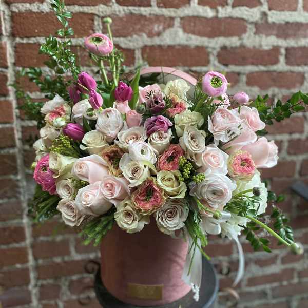 Round hatbox arrangement of blush roses, pink ranunculus, and tulips in a pink container