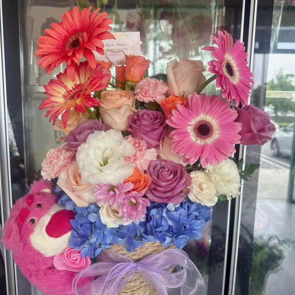 Mixed pink and purple flower arrangement with a plush bear and ribbon