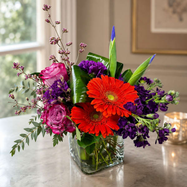 Bright mixed bouquet with orange gerbera daisies, pink roses, and purple flowers in a glass vase