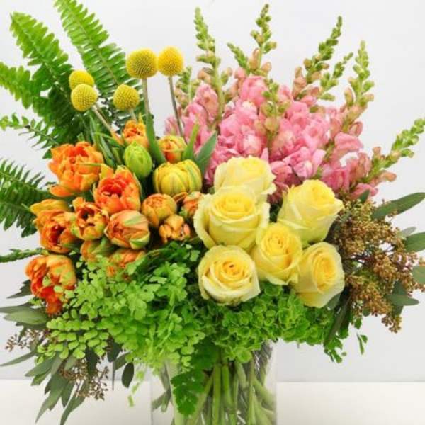 Bright arrangement of yellow roses, orange tulips, pink snapdragons, and ferns in a clear glass vase