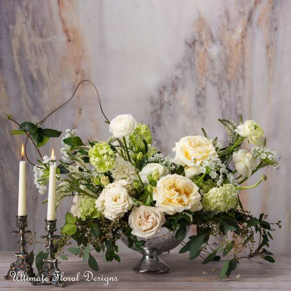 Cream and white floral arrangement in a silver bowl beside two lit candles