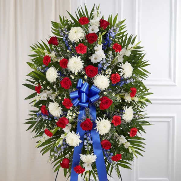 Patriotic standing spray with red roses, white mums, blue delphinium and a large blue ribbon on an easel.