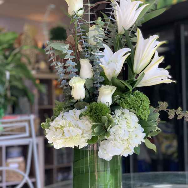 Tall white arrangement with lilies, roses, and hydrangeas in a clear green-lined glass vase.