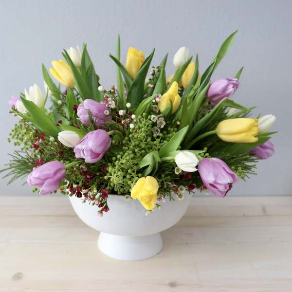 Low arrangement of yellow, white, and lavender tulips in a white pedestal bowl