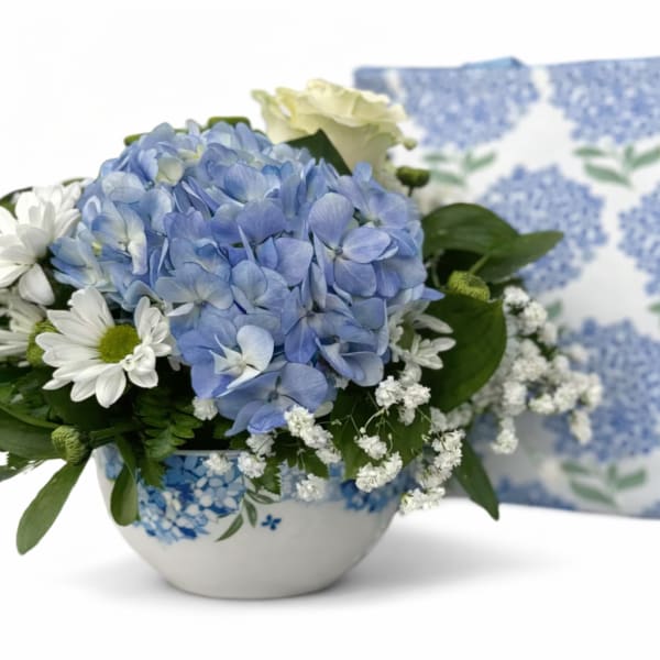 Blue hydrangea bouquet in a white ceramic bowl with a matching patterned pillow behind it