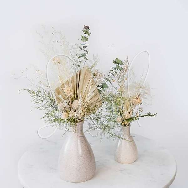 Two dried floral arrangements in cream vases on a round table