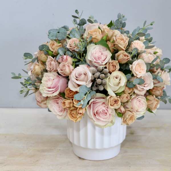 Round arrangement of blush and peach roses in a white ceramic vase with soft gray pods