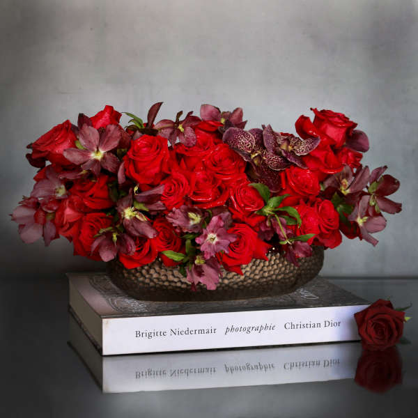 Low arrangement of red roses and burgundy flowers in a metal bowl displayed on stacked books.