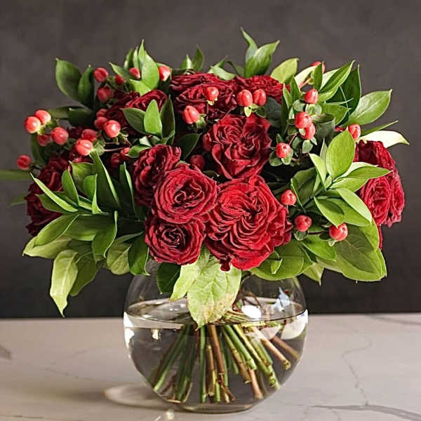 Red roses in a clear glass vase with green foliage and red berries