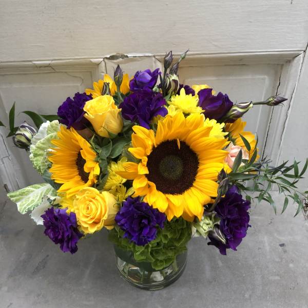 Bouquet of sunflowers, yellow roses, and purple blooms in a glass vase