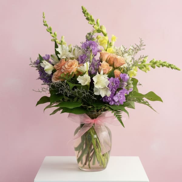 Mixed bouquet of purple, peach, and white flowers in a glass vase with a pink ribbon.