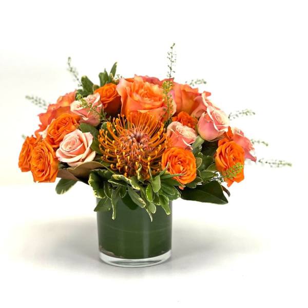 Low arrangement of orange and peach roses with a pincushion protea in a clear glass vase