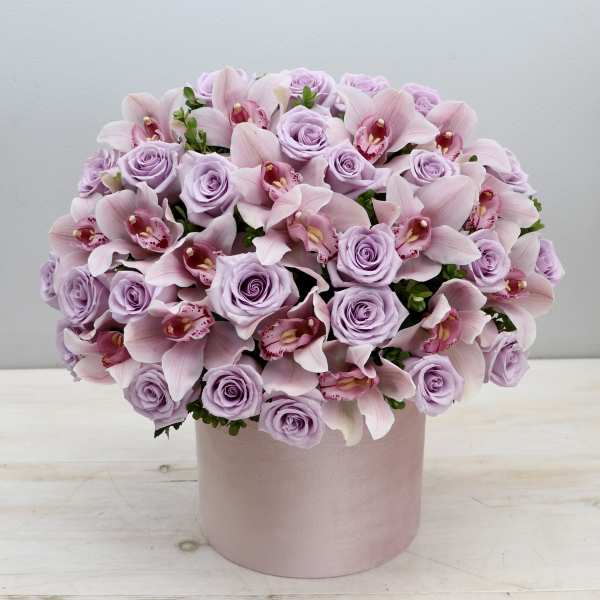 Round arrangement of pale lavender roses and pink orchids in a light pink hat box container.