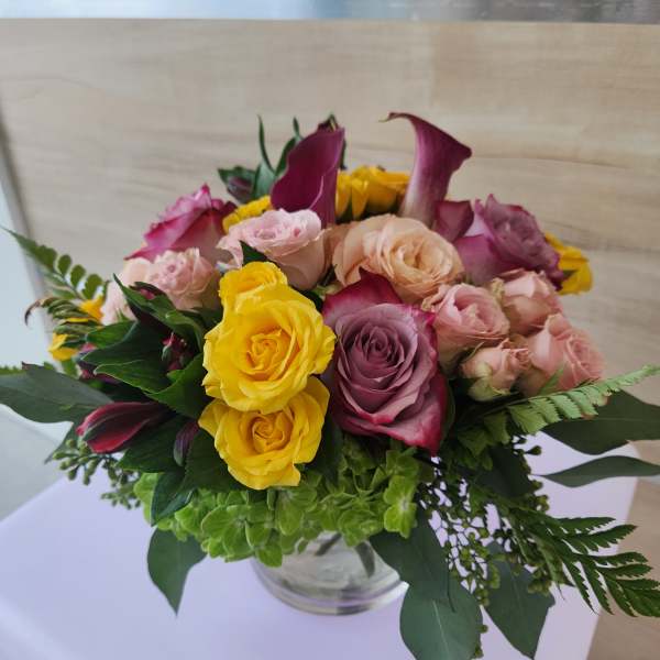 Round arrangement of yellow and pink roses with purple calla lilies in a clear glass vase