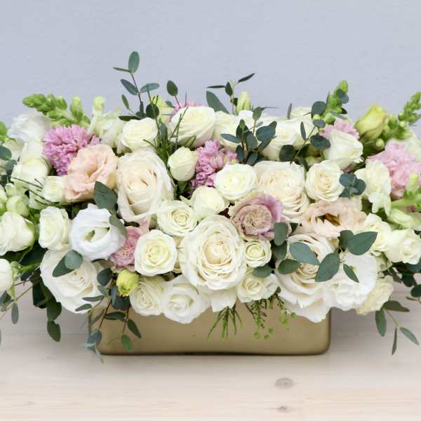 Low centerpiece of white and blush roses with green snapdragons in a gold rectangular vase