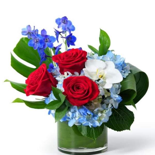 Short arrangement of red roses, blue flowers, blue hydrangea and a white orchid in a glass vase