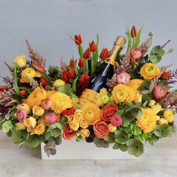 Rectangular arrangement of yellow and orange flowers with red tulips surrounding a bottle of champagne.