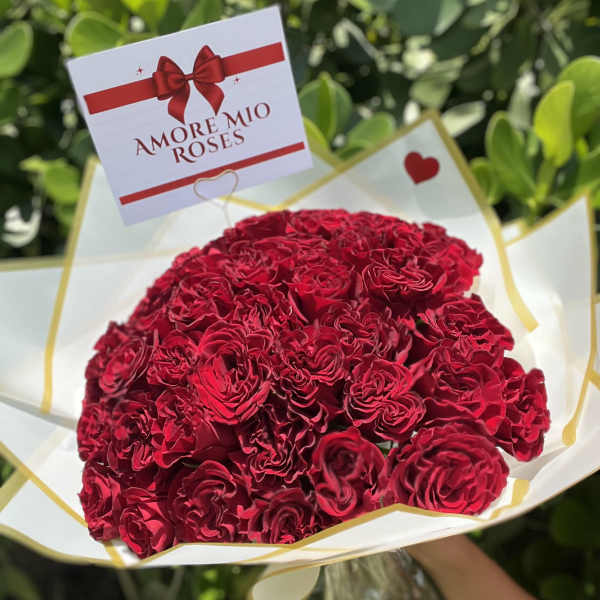 Hand-tied bouquet of tightly clustered red roses wrapped in white paper with an Amore Mio Roses card.