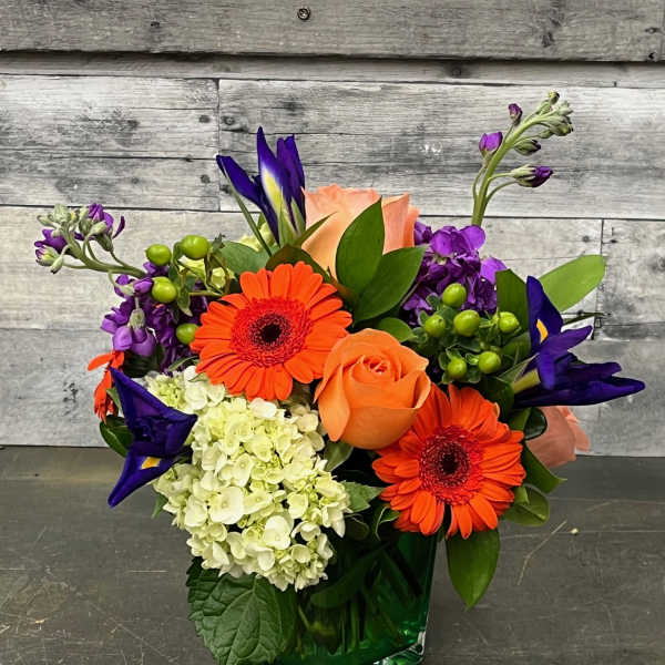 Bright mixed bouquet with orange gerberas, peach roses, and purple blooms in a green vase.